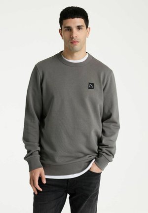 Joven con sudadera gris con un pequeño logo en el pecho, camiseta blanca y pantalones negros, de pie frente a fondo blanco.