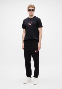 Μαύρο βαμβακερό μπλουζάκι με λογότυπο, σε συνδυασμό με μαύρες φόρμες με λογότυπο και λευκά sneakers, σε απλό φόντο.