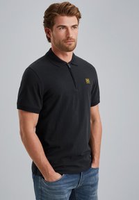 Pánské polo tričko z černé bavlny s límečkem na zip. Na hrudi je malé žluté logo a krátké rukávy. Je oblečeno s modrými džíny.
