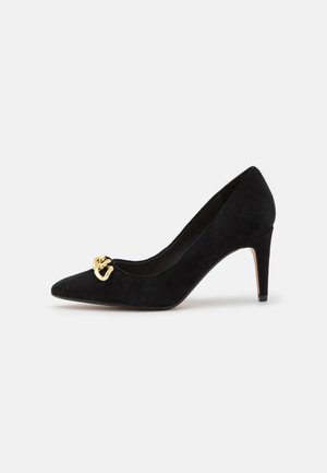 Zapato de tacón alto de ante negro con punta afilada y un detalle de cadena dorada en el empeine. Textura suave, tacón de aproximadamente 7,5 cm.