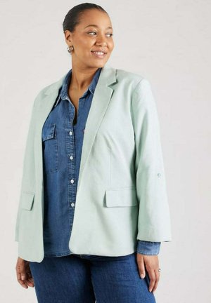 Blazer verde menta chiaro realizzato in tessuto morbido, con un design sartoriale, due tasche frontali e una vestibilità leggermente rilassata, indossato sopra una camicia di denim.