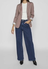 Kobieta w fioletowym żakiecie z podwiniętymi rękawami, białej bluzce, szerokich niebieskich dżinsach z wysokim stanem i czarnych, spiczastych botkach, pozująca z jedną ręką w kieszeni.