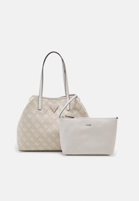 Sac fourre-tout tissé beige avec longues anses à côté d'une petite pochette crème zippée avec logo Guess doré sur fond blanc uni.