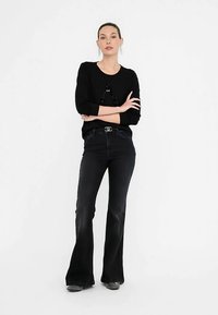 Črna majica z dolgimi rokavi in okroglim izrezom, okrašena z biserčki v obliki črk, kombinirana s temnimi zvončastimi kavbojkami in ravnimi čevlji.