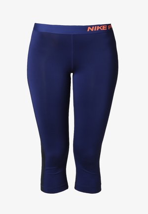 Frau geht nach vorne, trägt einen korallenfarbenen Sport-BH, marineblaue kurze Leggings und graue Laufschuhe vor einem einfarbigen Hintergrund.