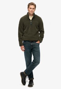 Zielono-oliwkowy polar z wysokim kołnierzem i pół zamkiem, zestawiony z niebieskimi dżinsami i czarnymi sznurowanymi butami. Gładka faktura, casualowy krój.