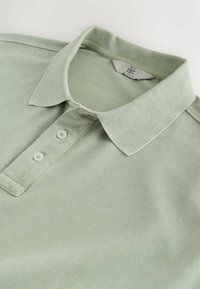 Polo vert clair avec deux boutons blancs et un col plié, posé à plat sur une surface blanche.