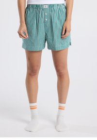 Vihreä- ja valkoruudulliset puuvillaiset bokserit, joissa on joustava vyötärönauha, nappikiinnitys ja sivuhalkiot, käytettynä valkoisten raidallisten sukkien kanssa.