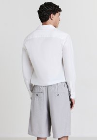 Homme de dos portant une chemise blanche à manches longues et un short gris clair à mi-cuisse avec des poches arrière boutonnées.