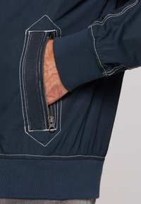 Navy blue jacket featuring a zippered pocket with herringbone fabric detail and white contrast stitching; ribbed cuffs and waistband.