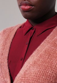 Lyserød tekstureret cardigan over en mørkerød skjorte med krave, med knapper og glat stof. Nærbillede af tøjets detaljer.