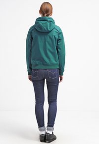 Grøn hættejakke med en glat, mat overflade, parret med mørkeblå skinny jeans og sorte sneakers med hvide såler.