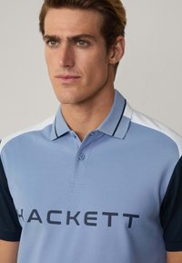 Polo di colore azzurro chiaro con maniche blu scuro, dotato di un'apertura con due bottoni e del logo "HACKETT" ben visibile sul davanti.