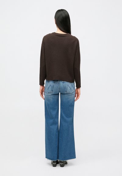 Rjav skrajšan pulover z okrolim izrezom, kombiniran s svetlo modrimi kavbojkami širokih hlačnic, ki imajo bleden vzorec in raztrgane robove.