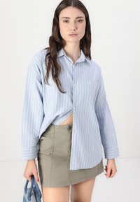 Camicia a righe blu chiaro con design a bottoni, abbinata a una mini gonna khaki. La camicia presenta una tasca, un tessuto morbido e maniche lunghe.