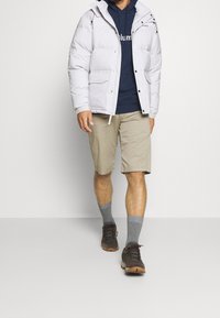 Manligt bärande en ljusgrå pufferjacka över en marinblå hoodie, beiga shorts, grå strumpor och bruna vandringsskor som går framåt.