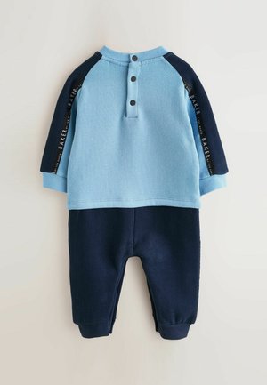 Light blue and navy romper with long sleeves, button placket at the back, and text detail along the shoulder seams. Soft fabric texture.