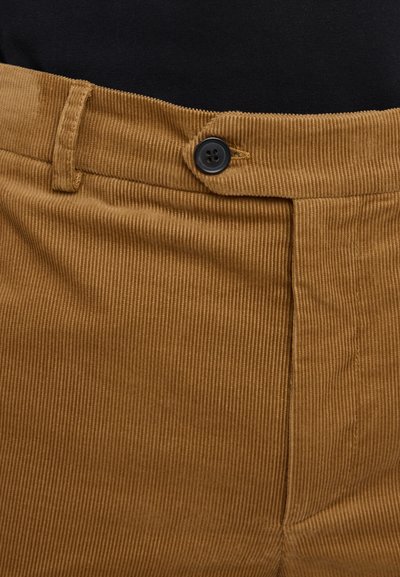 Un pantalon en velours côtelé marron présente une finition texturée, un motif à côtes verticales et un bouton noir de fermeture au niveau de la taille.