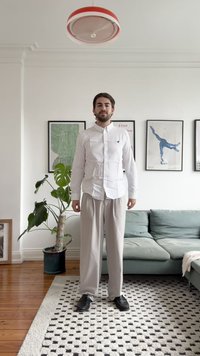 Un bărbat stă într-un interior luminos, purtând o cămașă albă cu nasturi și pantaloni gri deschis, cu pantofi negri. O plantă și o canapea se află în apropiere.