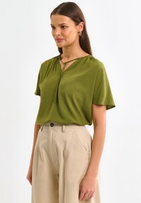 Alyvučių žalios spalvos trumpomis rankovėmis bluzonė su raktų skylutės iškirptės raišteliu; švelni tekstūra ir laisvas kirpimas, derinama su šviesiai smėlio spalvos aukštu liemeniu kelnių.