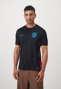 Trumpo rankovėmis juodas sportinis marškinėlis su mėlynu Nike logotipu ir Anglijos emblemą; ant šono yra ryškus gradientinis raštas, suteikiantis vizualų kontrastą.