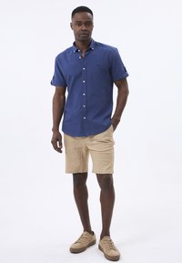 Camisa de manga corta azul con botones en la parte delantera, combinada con pantalones cortos beige y zapatillas color beige. La camisa tiene cuello y un detalle de logo sutil.