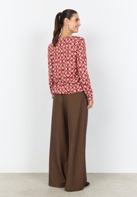 Langärmliges Oberteil mit rotem und cremefarbenem Muster, abgerundetem Ausschnitt, kombiniert mit braunen Hosen mit weitem Bein. Strukturierter Stoff, legerer Schnitt.