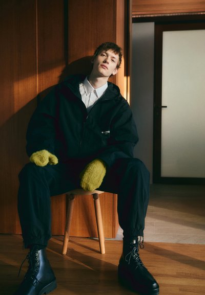 Black windbreaker with hood, white shirt, green knitted mittens, black pants, and chunky black lace-up boots, seated on a wooden stool.