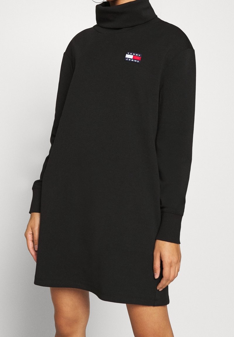 Robe sweat noire avec col montant, manches longues et petit logo brodé sur la poitrine. Tissu lisse avec une coupe décontractée.