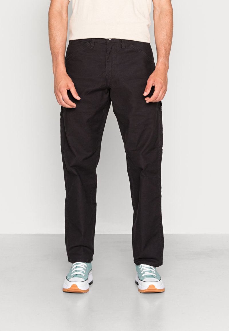 Man med svarta raka byxor och ljusblå sneakers med vita sulor som står på ett grått golv mot en vit bakgrund.