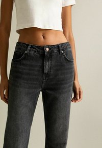Mujer vistiendo jeans negros desgastados de cintura alta y una blusa corta blanca de mangas cortas, de pie contra un fondo neutro claro.