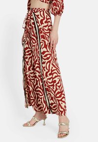 Falda maxi en crema con un patrón abstracto rojo, que presenta una cintura con cordón y franjas de acento en negro. Combinada con sandalias de tacón metálicas.