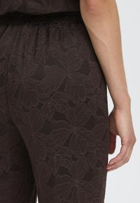 Rudas tekstūruotas audinys su gėlių raštu, turintis prigludusį liemenį ir subtilų blizgesį. Detalus drabužio nugaros vaizdas.