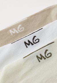 Tres pares de calcetines en beige, blanco y verde claro. Cada uno presenta las letras "MG" bordadas en colores contrastantes en el puño.