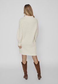 Robe-pull en maille crème à manches longues avec texture côtelée, ornée de boutons décoratifs au niveau du poignet. Portée avec des bottes de cow-boy marron.