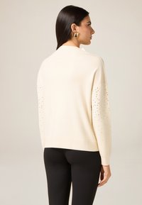Kremowy sweter z dzianiny o luźnym kroju, z ozdobnymi srebrnymi ćwiekami na rękawach. Gładka tekstura i ściągacz na dole.