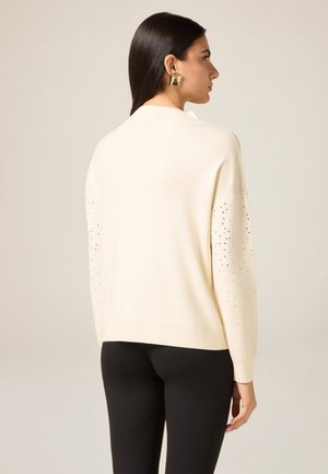 Kremowy sweter z dzianiny o luźnym kroju, z ozdobnymi srebrnymi ćwiekami na rękawach. Gładka tekstura i ściągacz na dole.