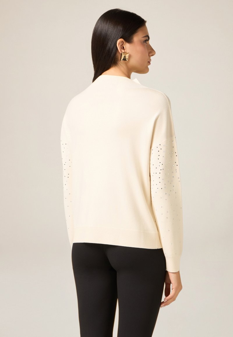 Kremowy sweter z dzianiny o luźnym kroju, z ozdobnymi srebrnymi ćwiekami na rękawach. Gładka tekstura i ściągacz na dole.