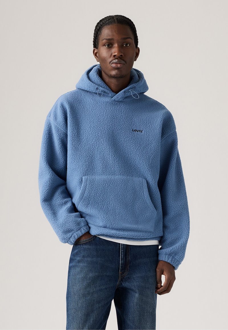 Hoddie en polaire bleu clair avec une surface texturée, poche frontale et logo "Levi's" noir. Manches longues avec des bords côtes. Porté avec un jean foncé.
