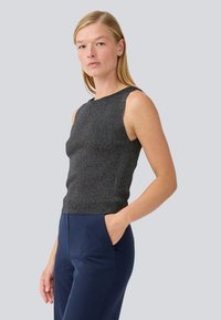 Sleeveless top in dark gray with a subtle shimmer, featuring a fitted design and scoop neckline. Paired with navy trousers, hands in pockets.
