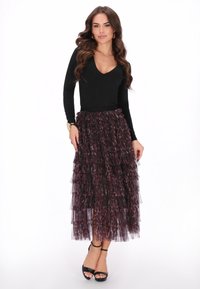 Femeie cu păr lung și ondulat, purtând un top negru, ajustat, cu decolteu în V, o fustă din tul cu model purpuriu închris, cu volane, pantofi cu toc la gleznă negri și o brățară aurie.