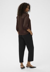 Blusa de satén marrón con mangas tres cuartos y dobladillo redondeado, combinada con pantalones negros ajustados y tacones tipo slingback.