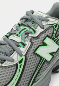 Pelēka sporta apava ar tīkla augšdaļu, kurā ir zaļas akcenti, teksturētas auklas un balts logotips. Dizainu izceļ melni elementi.