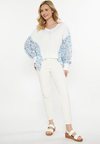 Top bianco a maglia con maniche a fantasia floreale blu, scollatura a V e polsini arricciati, abbinato a pantaloni slim-fit crema e sandali con zeppa marroni.
