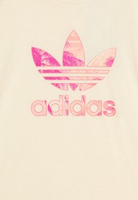Kremkrāsas T-krekls ar lielu rozā Adidas logotipu, kurā ir lapu dizains un diognālas svītras, izgatavots no mīksta kokvilnas materiāla.