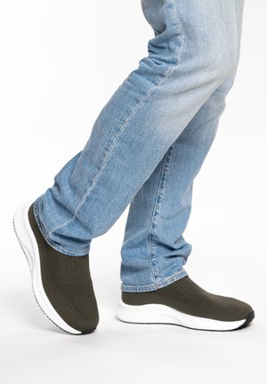 Beine in hellblauen Jeans und dunkelolivgrünen, gestrickten Slip-On-Sneakern mit dicken weißen Sohlen.