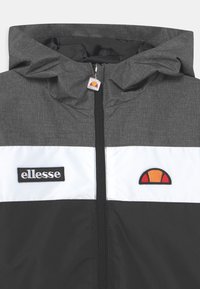 Giacca grigia con cappuccio, caratterizzata da una striscia bianca e dal logo nero "ellesse". Include un'emblema arancione e rosso. Tessuto testurizzato.