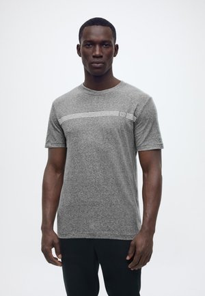 Άνδρας με κοντά μαλλιά που φοράει γκρι κοντομάνικη μπλούζα με λευκές οριζόντιες ρίγες και μαύρο παντελόνι, στέκεται μπροστά σε λευκό φόντο.