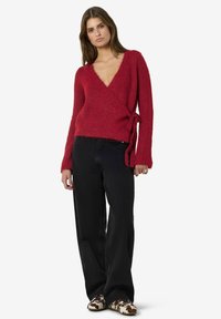 Kvinna som bär en djup röd omslagströja, svarta vida jeans och loafers med koskinnsmönster, stående mot en vit bakgrund.