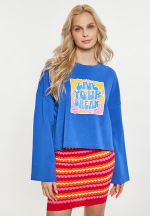 Ung kvinde med langt blondt hår iført en blå sweatshirt med teksten "Live Your Dream" og et farverigt stribet skørt.